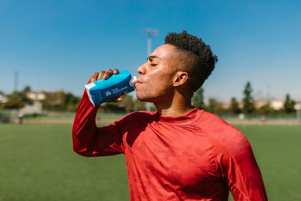 Adult athlete in a red shirt drinking water outdoors on a sunny day after sports activity.